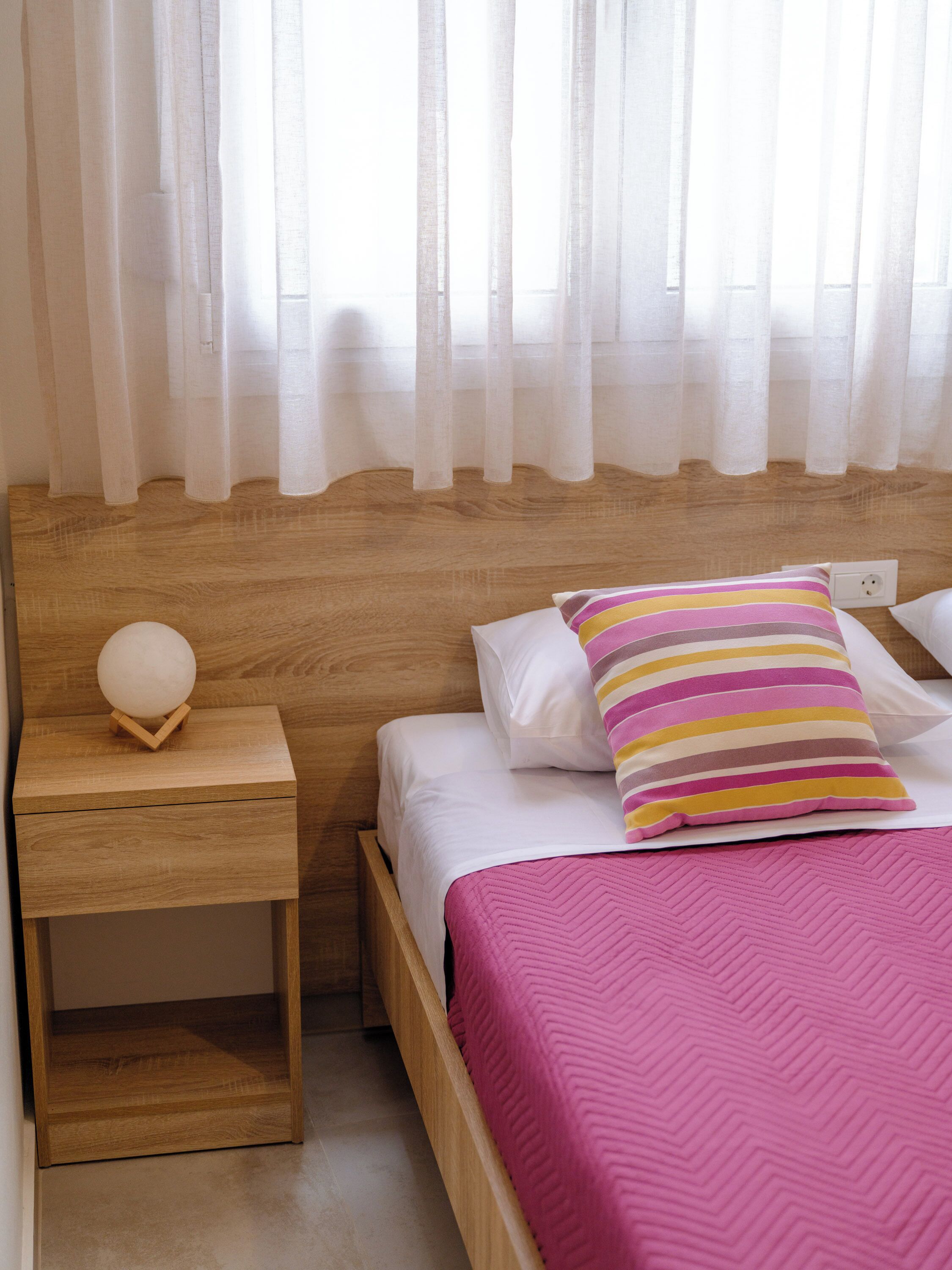 Un lit bien fait, recouvert d’une courtepointe rose, une table de chevet avec une lampe sphérique et des rideaux légers.