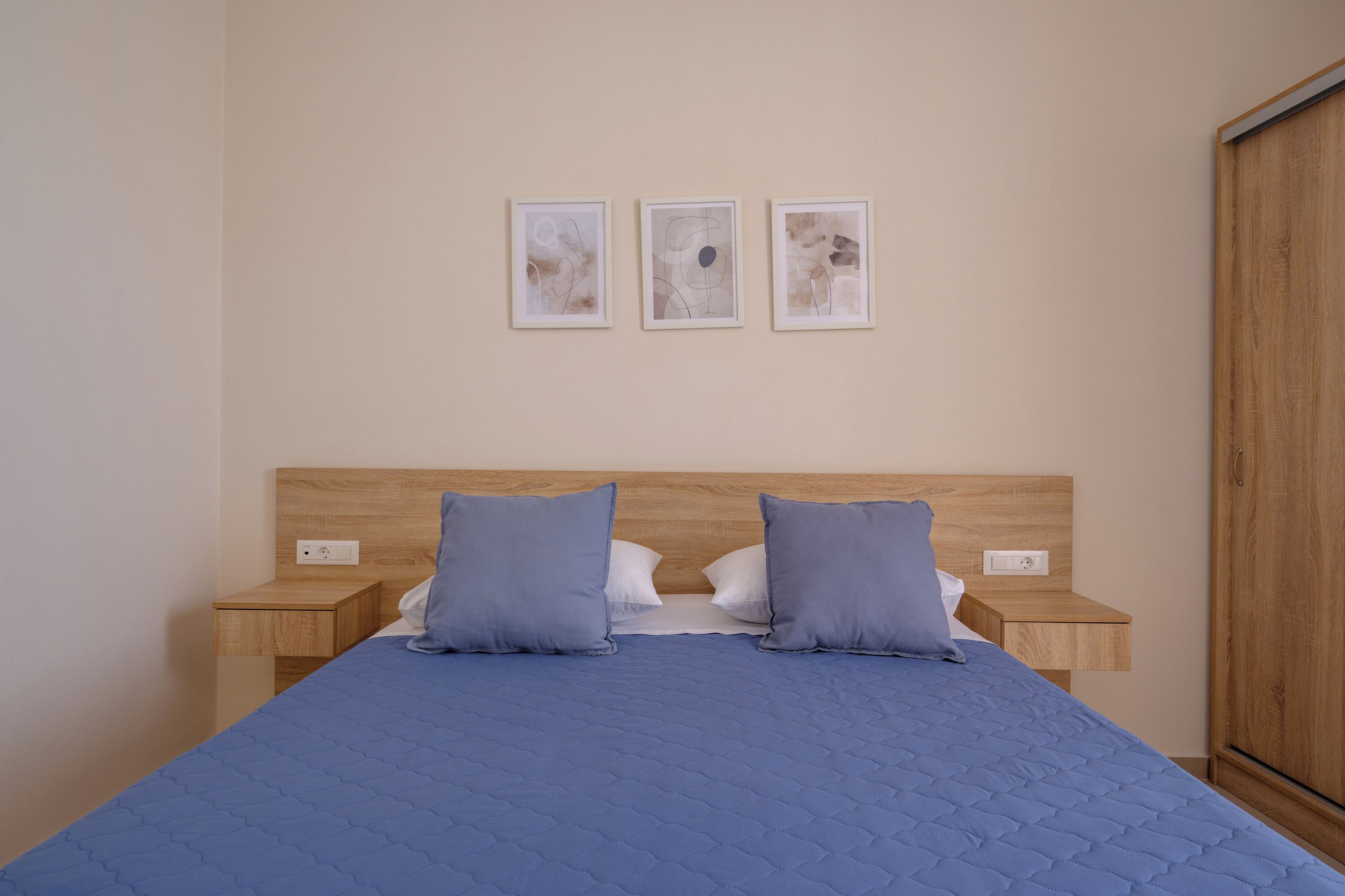 A neatly made bed with a blue quilt, two matching pillows, and a wooden headboard. There are three framed artworks on the wall above the bed.