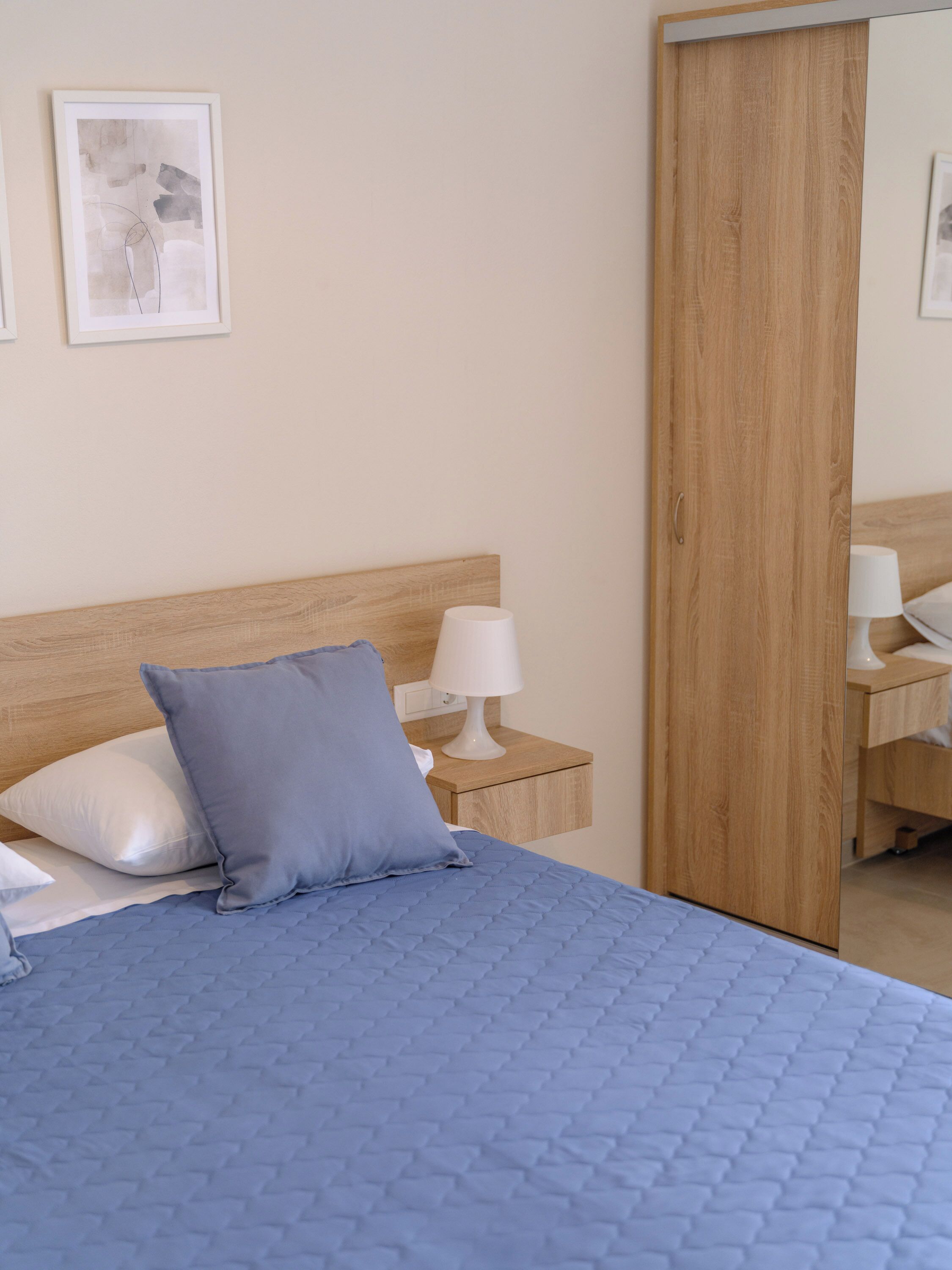 Un dormitorio con cabecera de madera, una cama con colcha azul, una mesita de noche con lámpara blanca y un armario de madera.