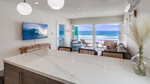 Una cocina moderna con una isla grande, un televisor en la pared y vistas a la playa a través de la puerta abierta.