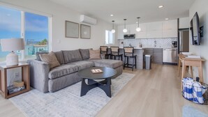 Un salon moderne avec un canapé d’angle gris, une table basse noire et un coin repas comprenant une table et des chaises en bois. On aperçoit à l’arrière-plan une cuisine avec des meubles blancs et des appareils électroménagers en acier inoxydable.