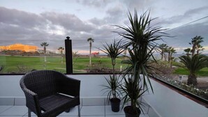 Uma varanda com cadeira de vime, plantas em vasos e vista para um campo de golfe e edifícios.