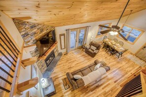 A spacious living area with a stone fireplace, wooden floors, a ceiling fan, and a dining area with a table and chairs. - Famtastico - Great Deal! Games, Hot Tub, Pets (Morganton)
