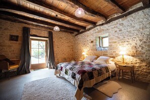 Une chambre à coucher rustique avec un mur en pierre, des poutres apparentes, un grand lit recouvert d’une courtepointe, une table de chevet avec une lampe et une fenêtre avec des rideaux.
