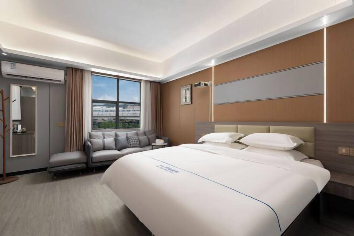 Quarto de hotel com uma cama grande, área de estar com sofá e poltrona, janela com vista e televisão na parede.