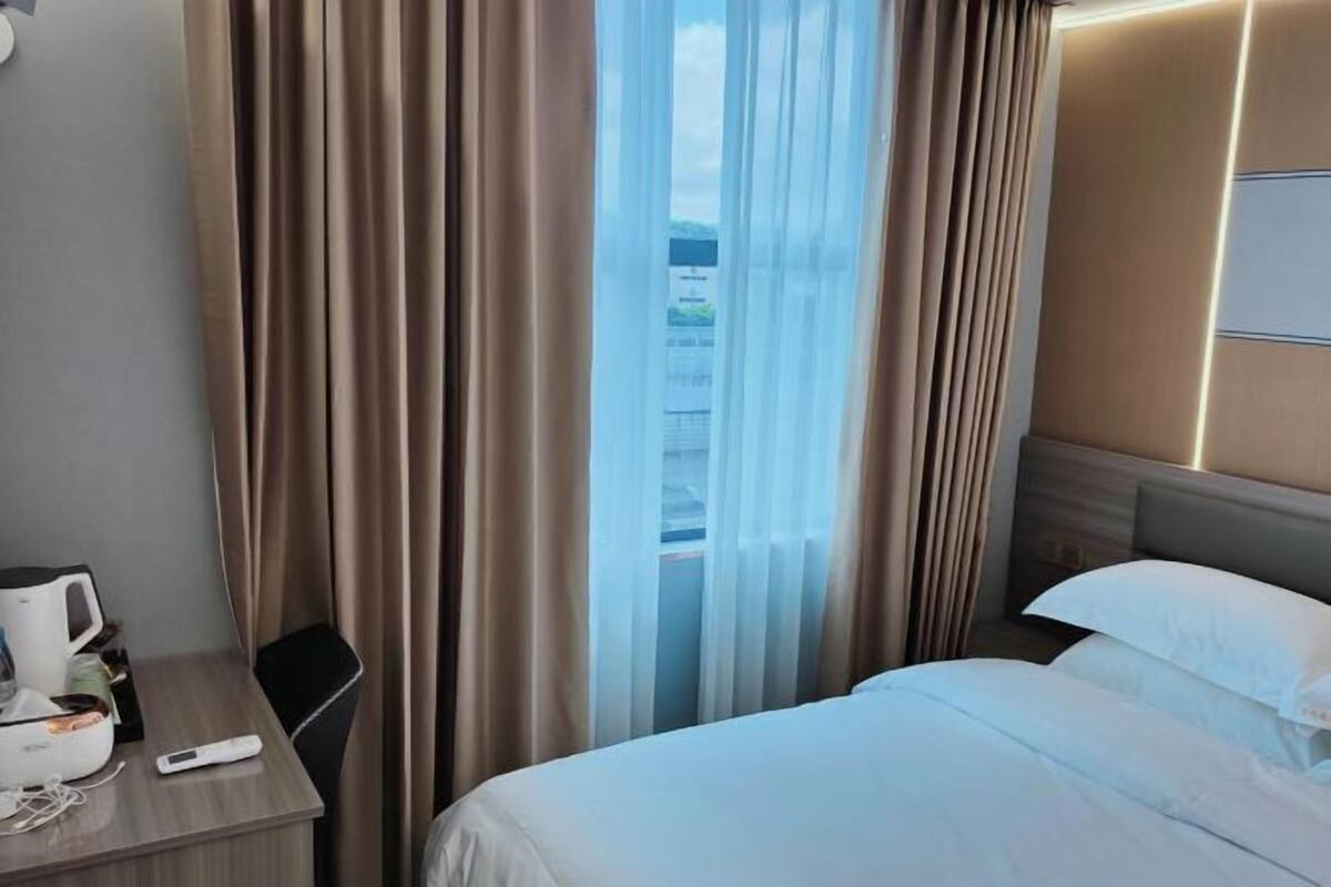 Quarto de hotel com cama, escrivaninha com cafeteira e janela com cortinas.