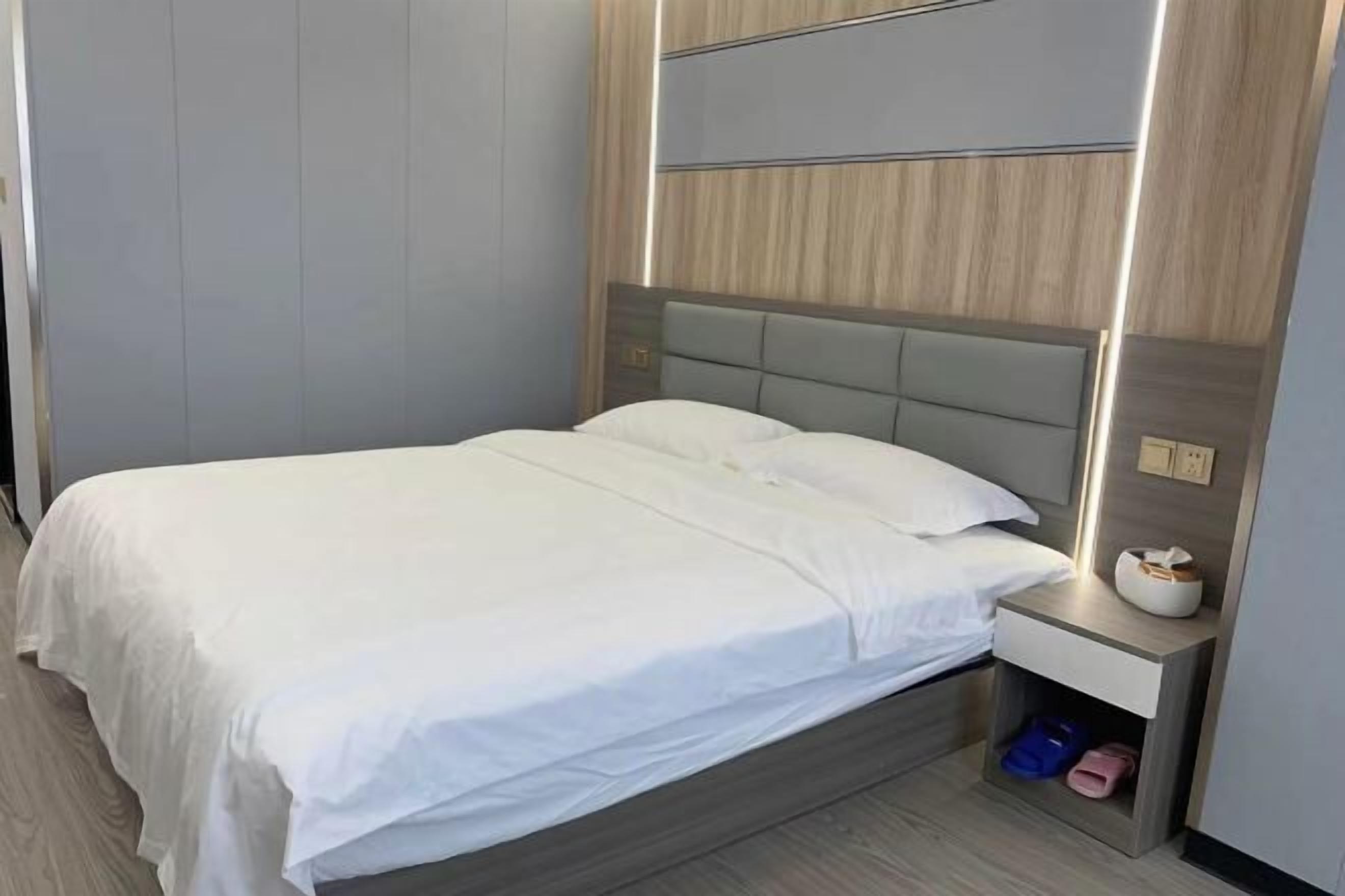 A neatly made bed with a headboard, a bedside table with a lamp, and a shoe rack under the bed.