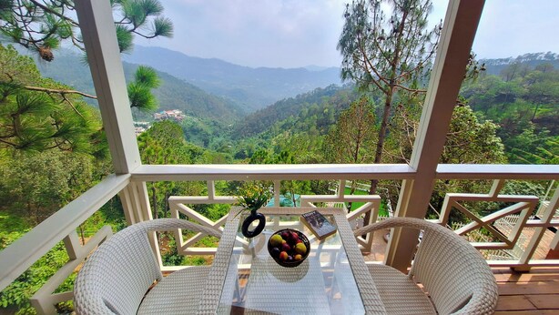 A balcony with wicker chairs and a table, overlooking a mountainous landscape. - Koro Treehouse Resort (Solan)