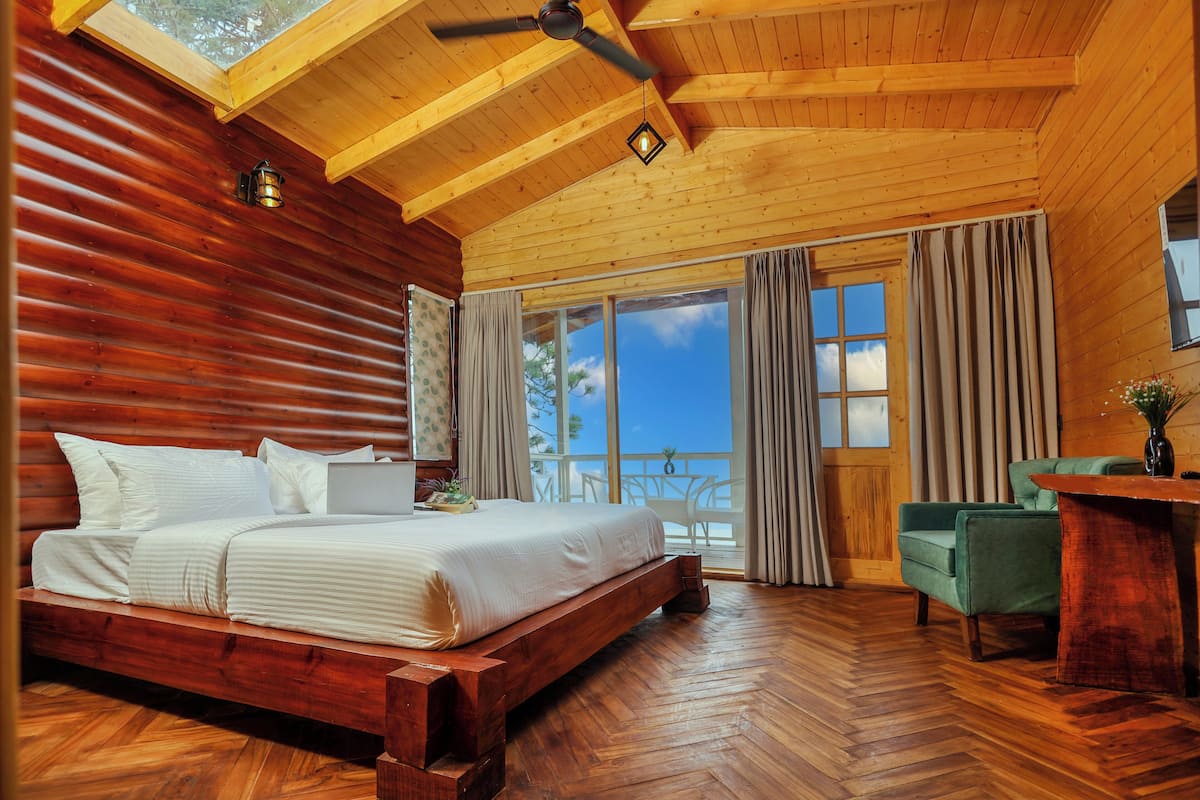 A bedroom with a wooden ceiling, a large bed, a desk, and a view of the outdoors.