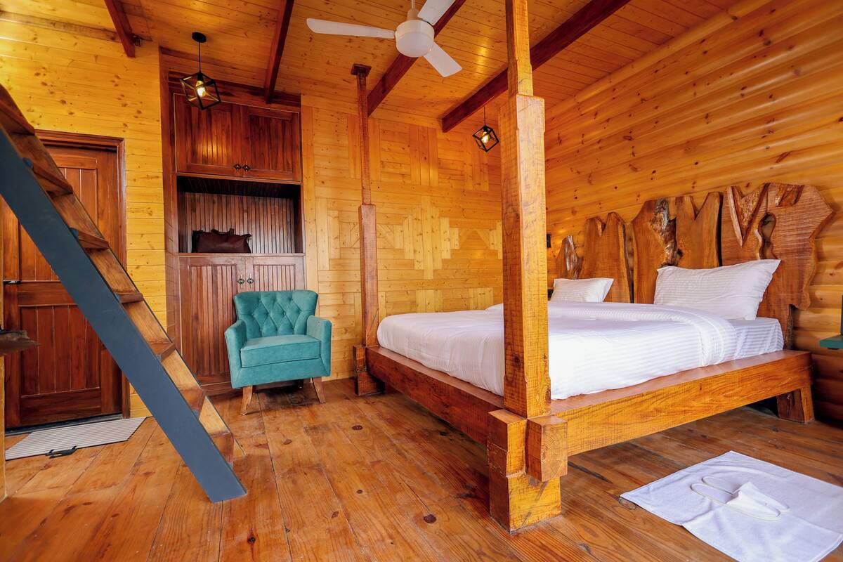 A wooden cabin room with a four-poster bed, a teal armchair, and a wooden wardrobe.