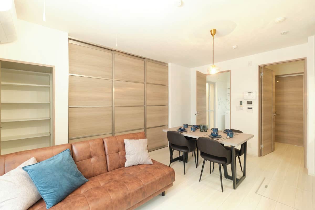 A modern living room with a sofa, dining table, and chairs. There is a large wardrobe and a door leading to another room.
