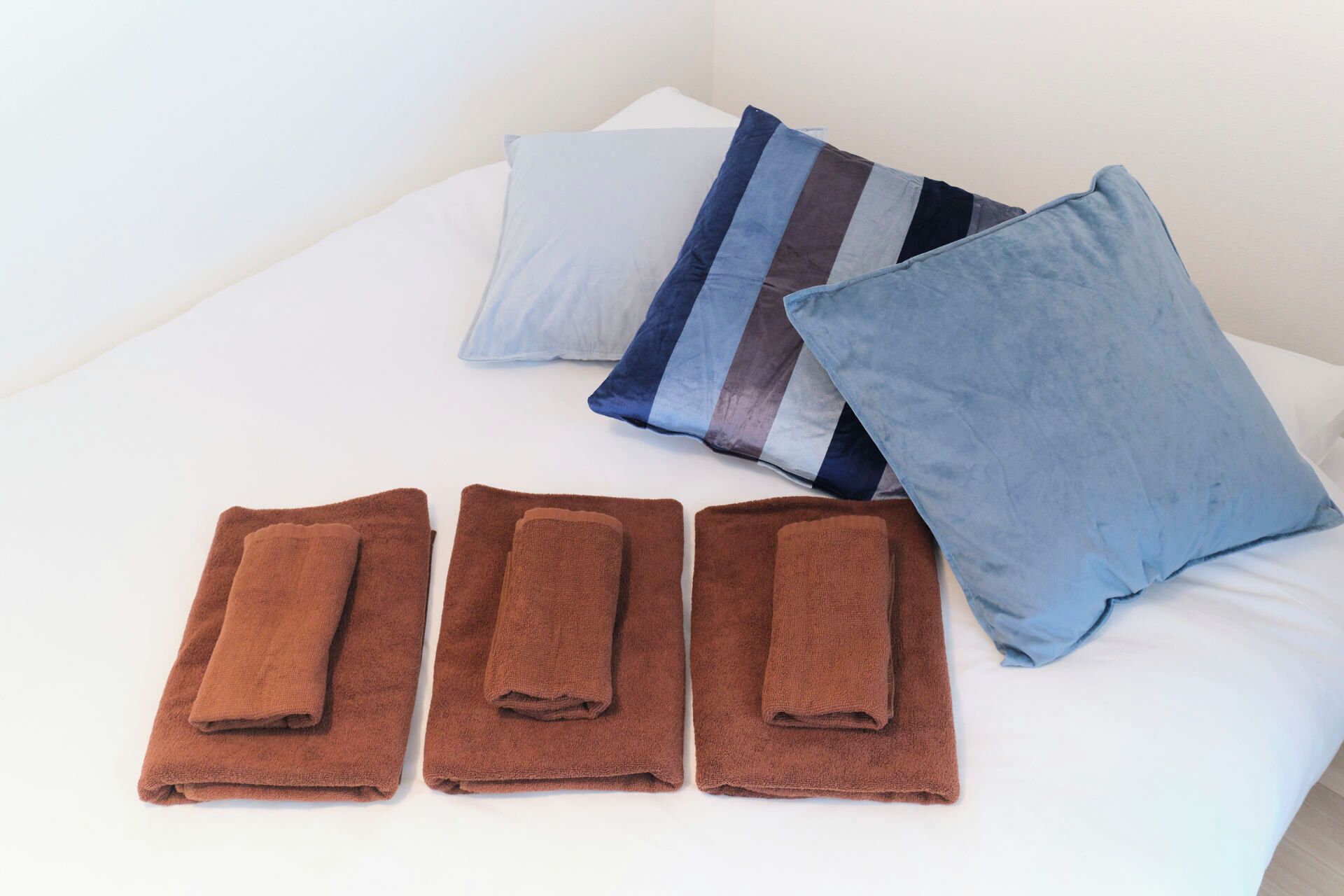 A bed with white sheets, three folded brown towels, and three pillows in blue, gray, and striped patterns.