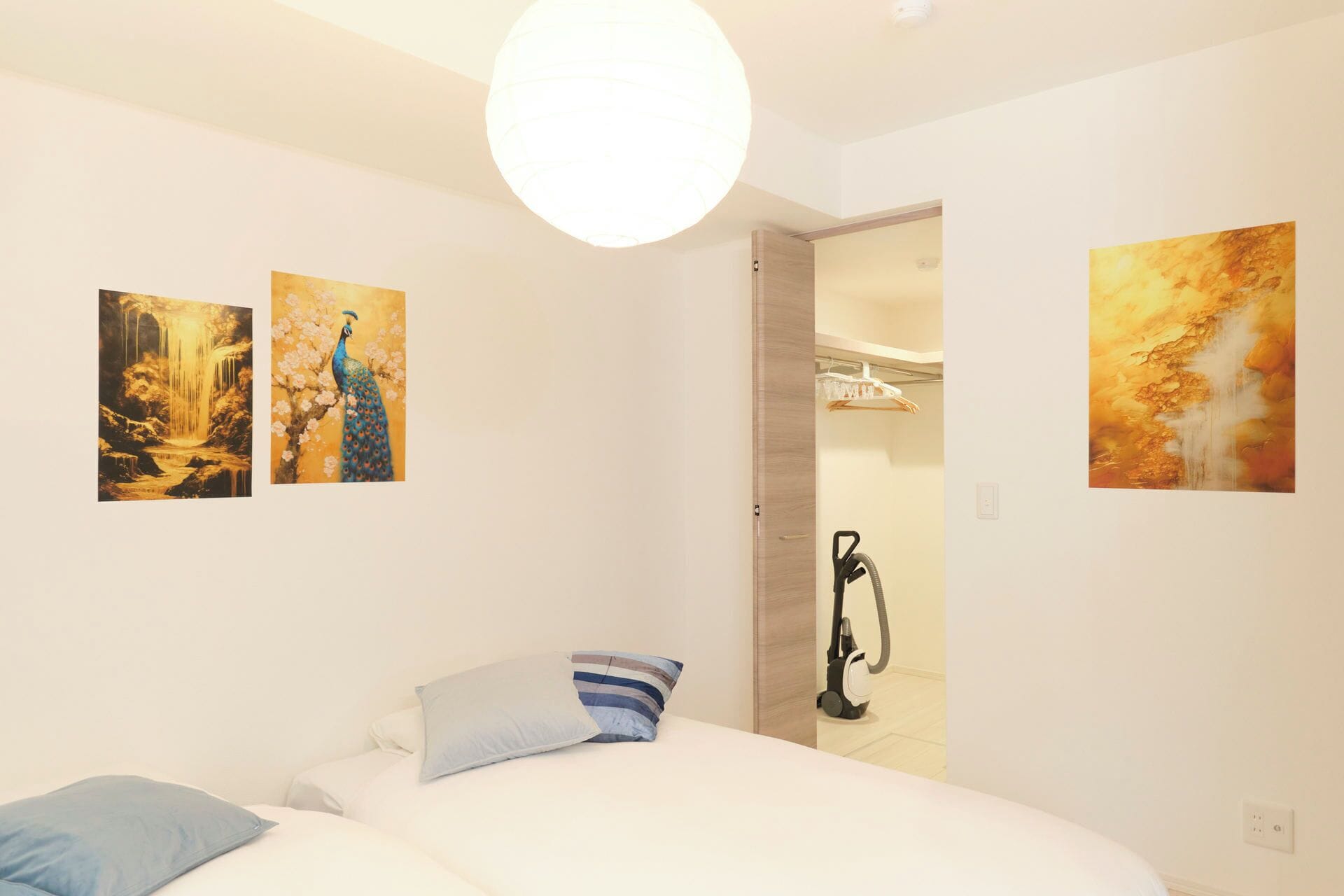 Quarto com cama, almofadas, um aspirador de pó e duas pinturas na parede.