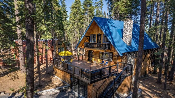 A cabin with a wooden deck, a stone fireplace, and a blue roof, surrounded by tall trees. - Modern Retro Tahoe Chic Chalet (Tahoe City)