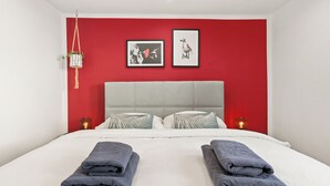 Ett hotellrum med en röd accentvägg, en säng med vita sängkläder, två kuddar och två hopvikta handdukar.