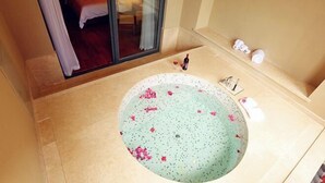 Un baño con una bañera grande, una puerta de vidrio que da a un dormitorio y un estante pequeño con artículos de tocador.