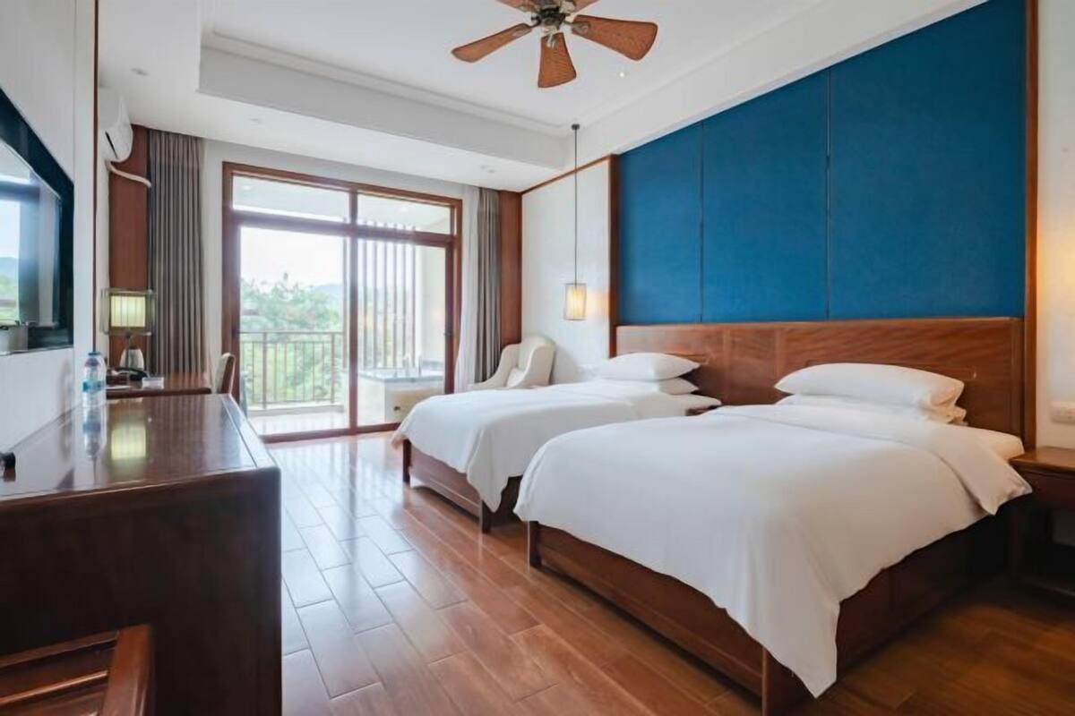 A hotel room with a desk, two beds, a ceiling fan, and a large window with a view.