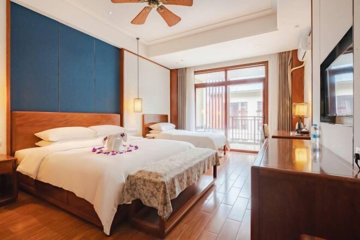 A hotel room with two beds, a desk, a ceiling fan, and a large window.