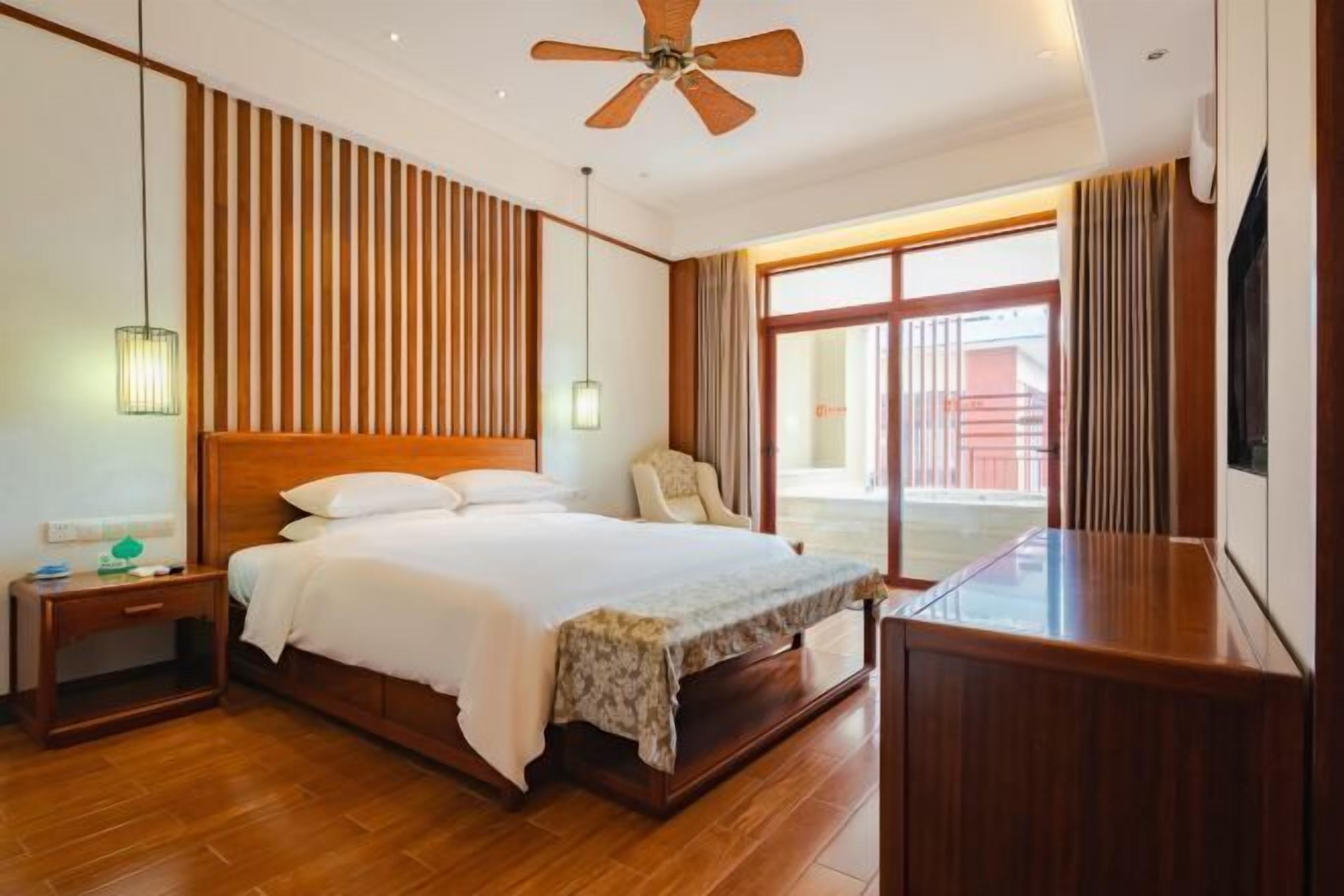 A hotel room with a wooden bed, a nightstand, a ceiling fan, a large window with curtains, and a TV on a wooden stand.