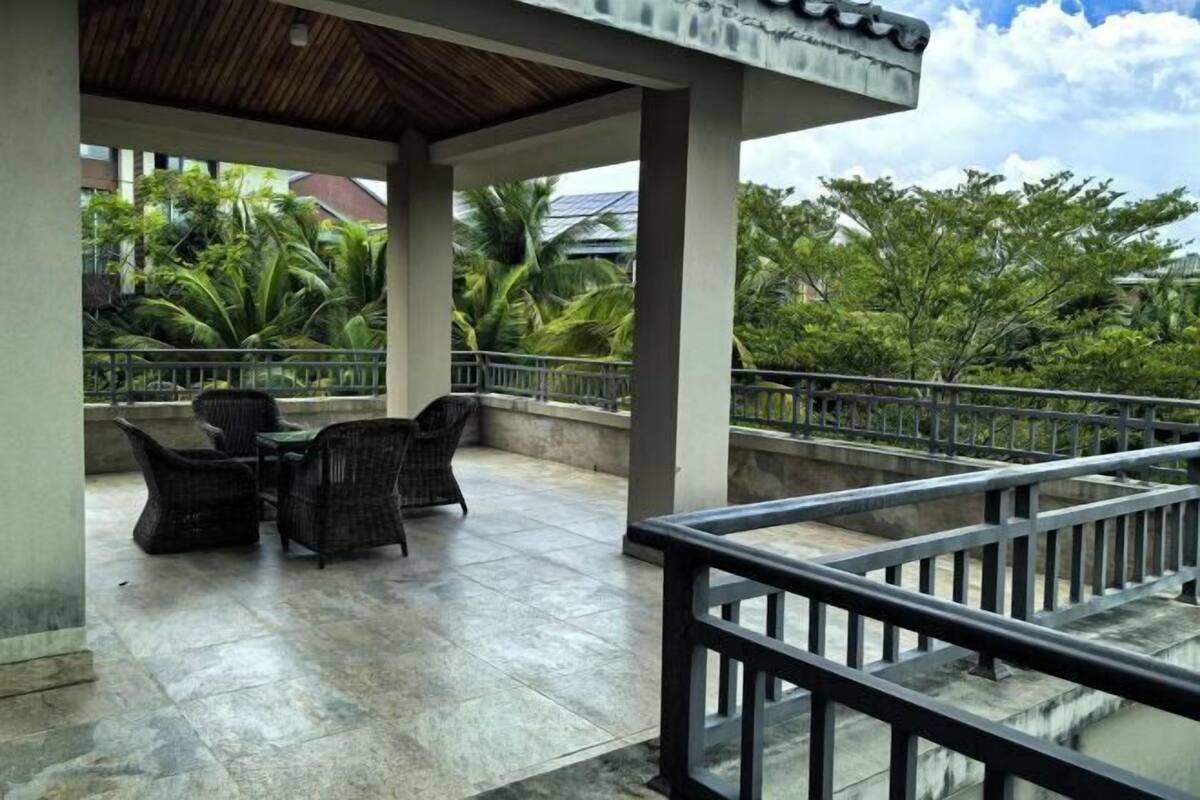 A covered outdoor seating area with wicker furniture and a tiled floor, overlooking a garden with palm trees.