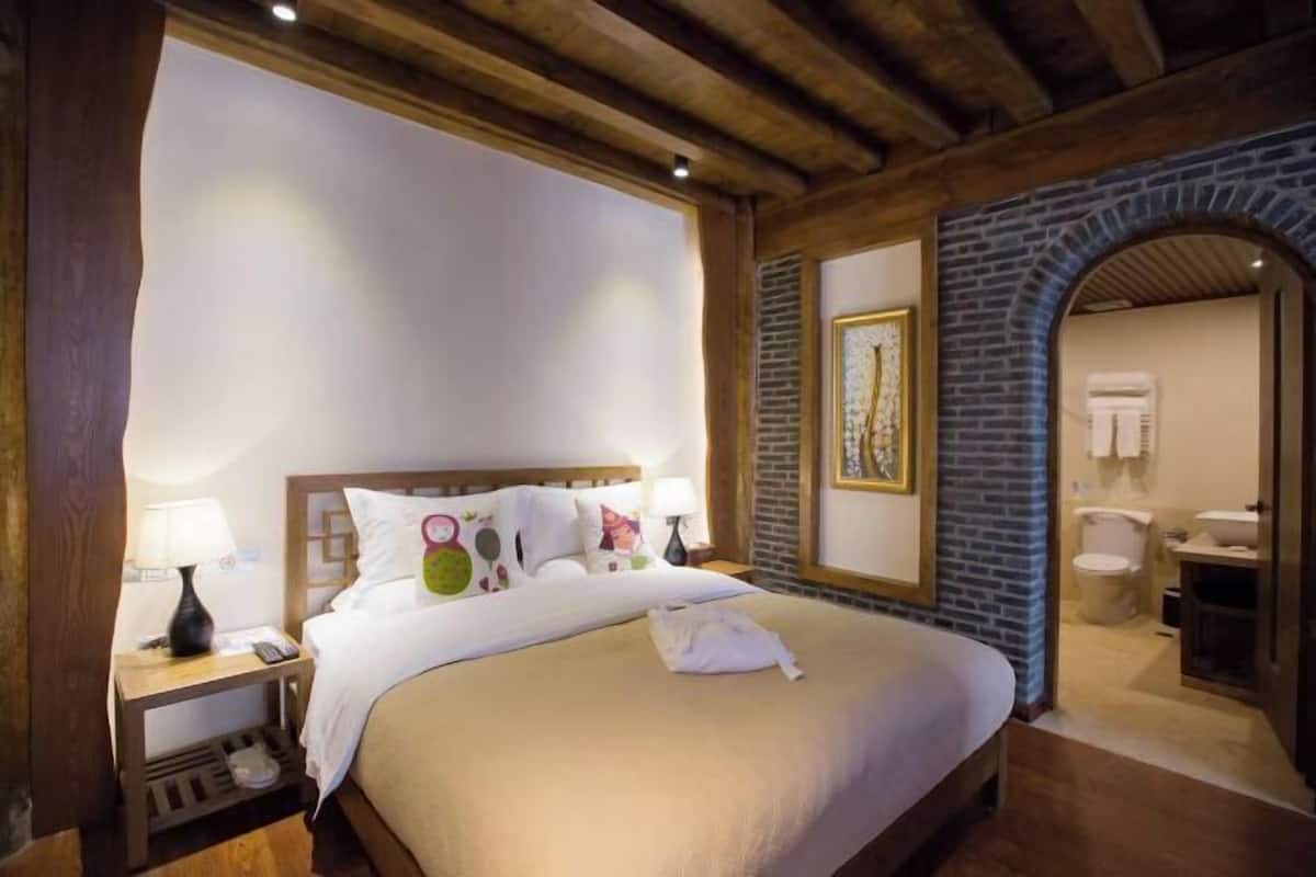 A hotel room with a wooden ceiling, a bed with white bedding, a nightstand with a lamp, and a bathroom visible through an archway.