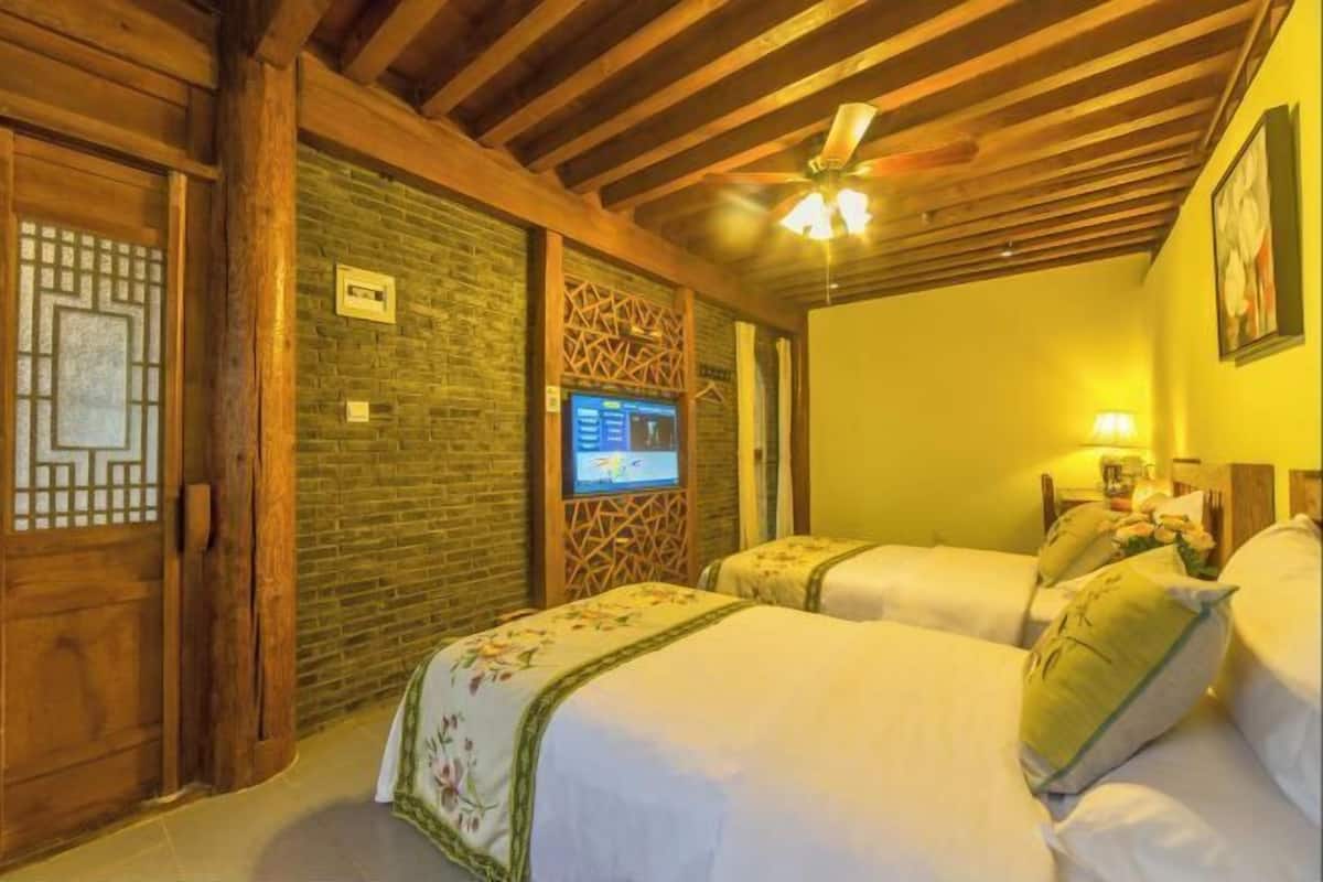 A hotel room with two beds, wooden beams, a brick wall, a ceiling fan, and a television on the wall.