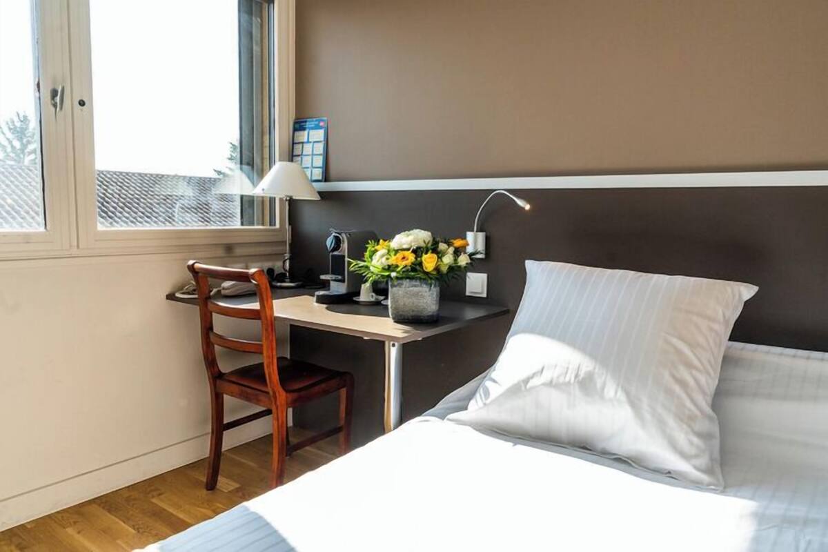 Une chambre d’hôtel comprenant un lit, un bureau, une chaise, une lampe et un arrangement floral.