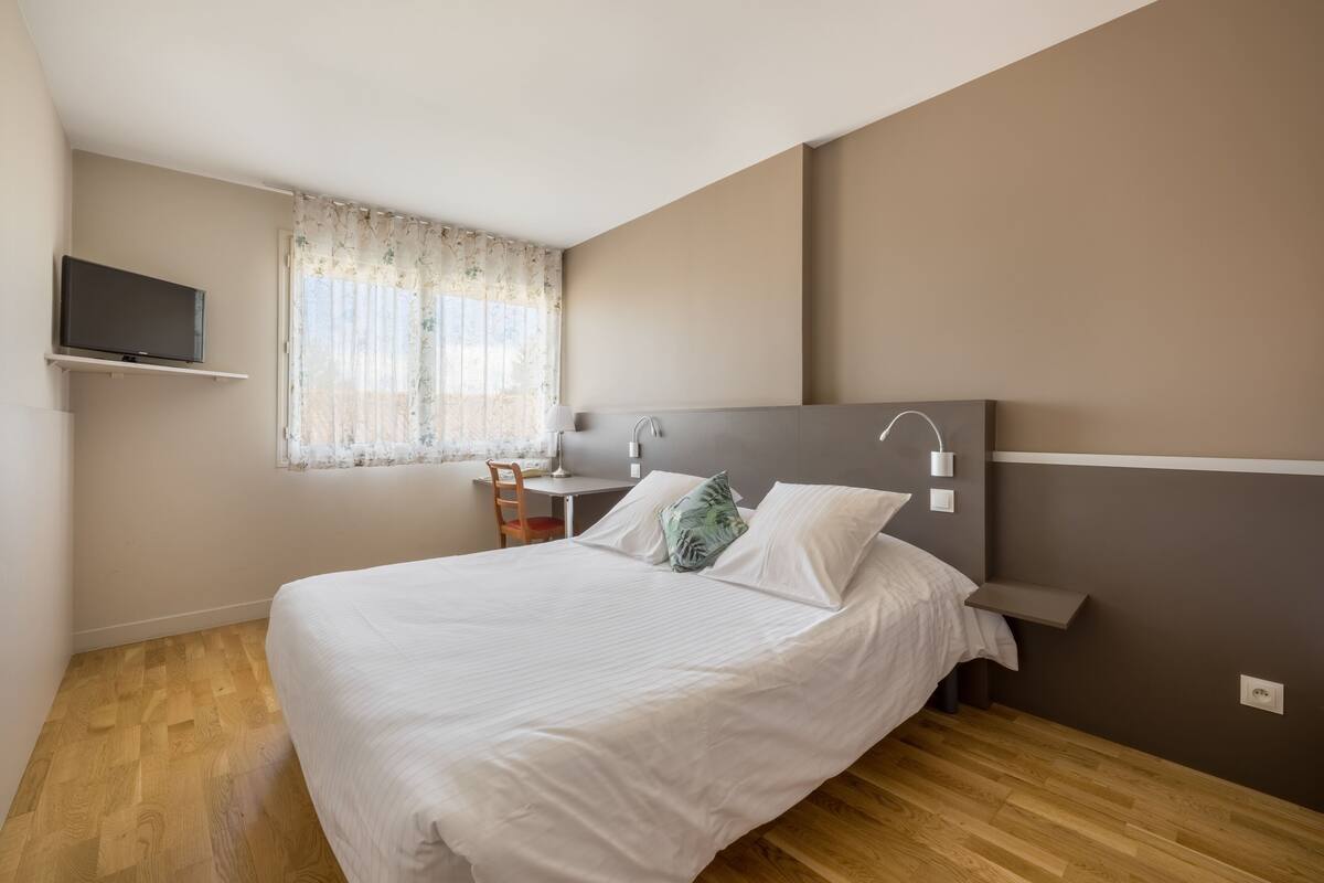 Une chambre à coucher avec un lit, un bureau, une chaise, une télévision et une fenêtre avec des rideaux.