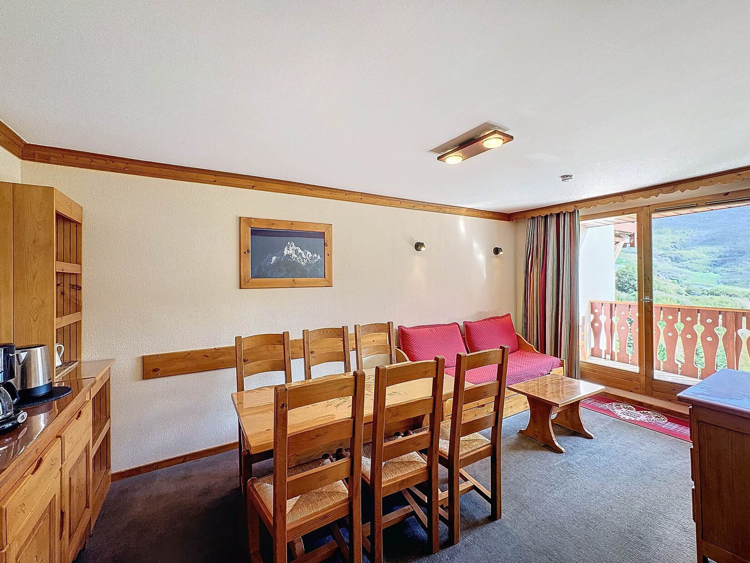 Une pièce chaleureuse avec une table à manger en bois, des chaises, un canapé avec des coussins rouges, un tableau encadré au mur et un balcon offrant une vue sur la campagne.