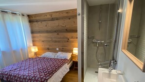 A bedroom with a wooden headboard, a bed with a red and white patterned bedspread, and a nightstand with a lamp. Adjacent is a bathroom with a glass shower enclosure, a white sink, and a mirror.