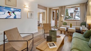 Ein modernes Wohnzimmer mit Holzboden, einem grünen Sofa, einem Couchtisch aus Holz und einem Sessel. An den Wänden hängen Bilder in Rahmen, und ein Fernseher zeigt eine Skiszenen.