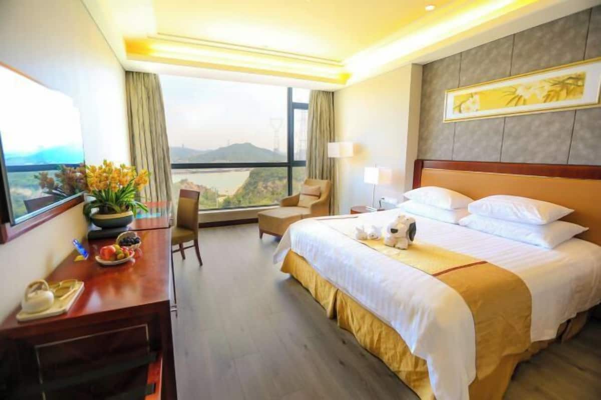A hotel room with a large bed, a desk with a vase of flowers, a chair, and a view of the cityscape.