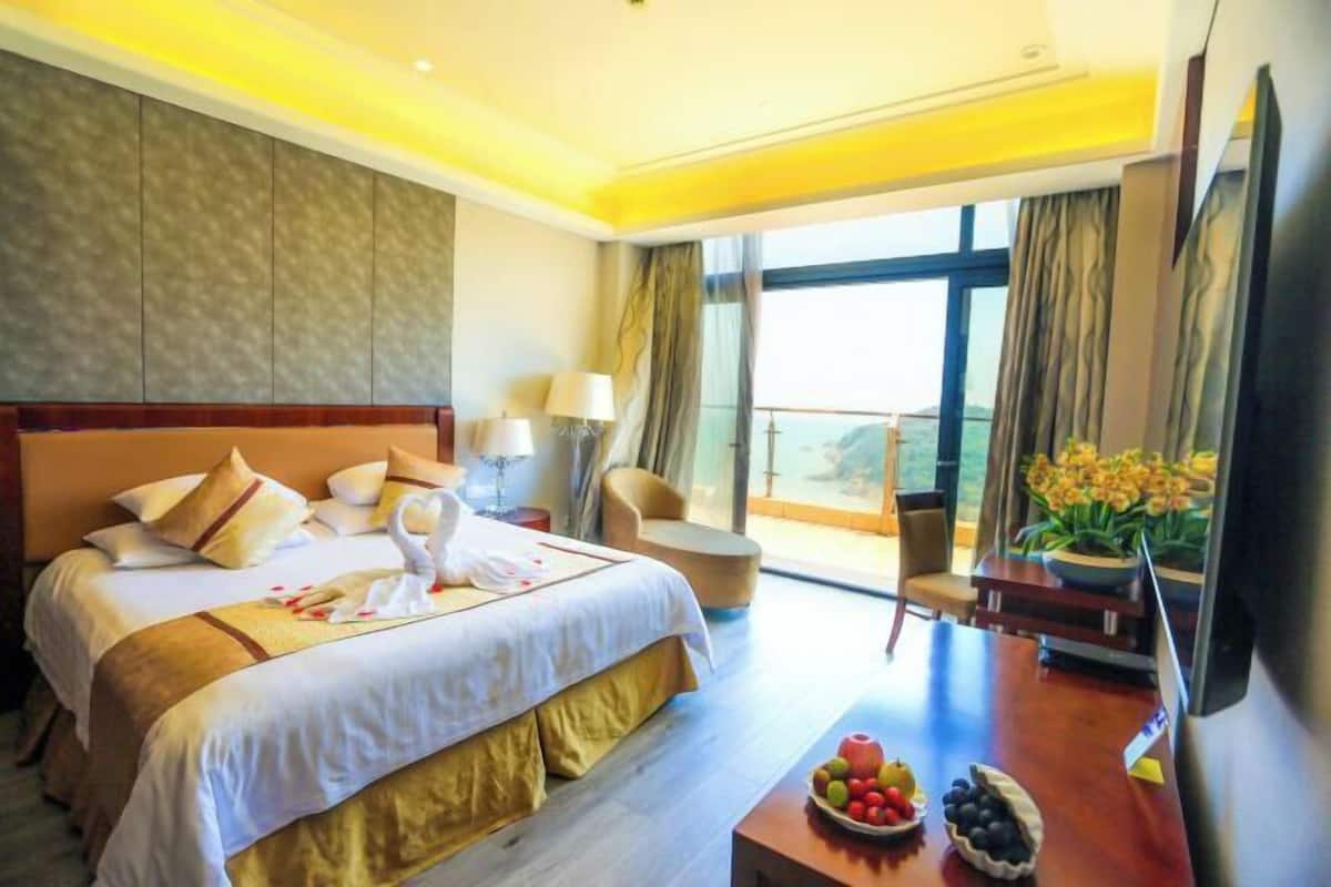A hotel room with a large bed, a desk with fruit, a chair, and a balcony with a view.