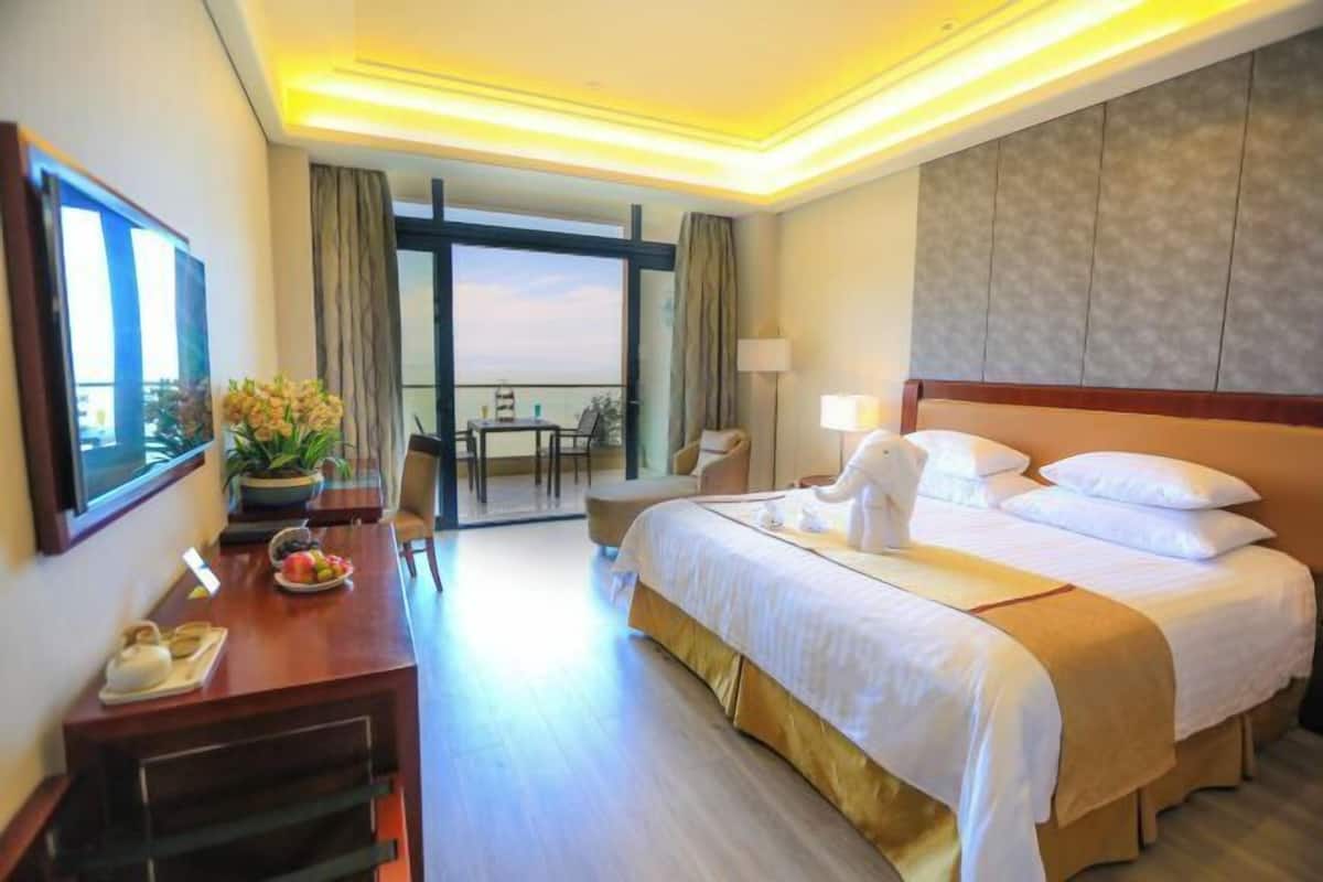A hotel room with a large bed, a desk with a telephone, a chair, a table with a plant, and a view of the outdoors.