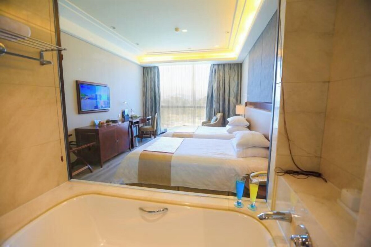 A hotel room with a large bathtub, two beds, a desk with a chair, a television, and a window with curtains.