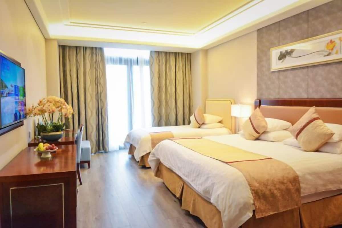 A hotel room with two beds, a desk with a vase of flowers, a television, and a painting on the wall.
