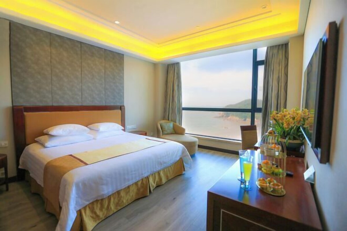 A hotel room with a large bed, a desk with a drink and snacks, a chair, and a view of the sea through a large window.