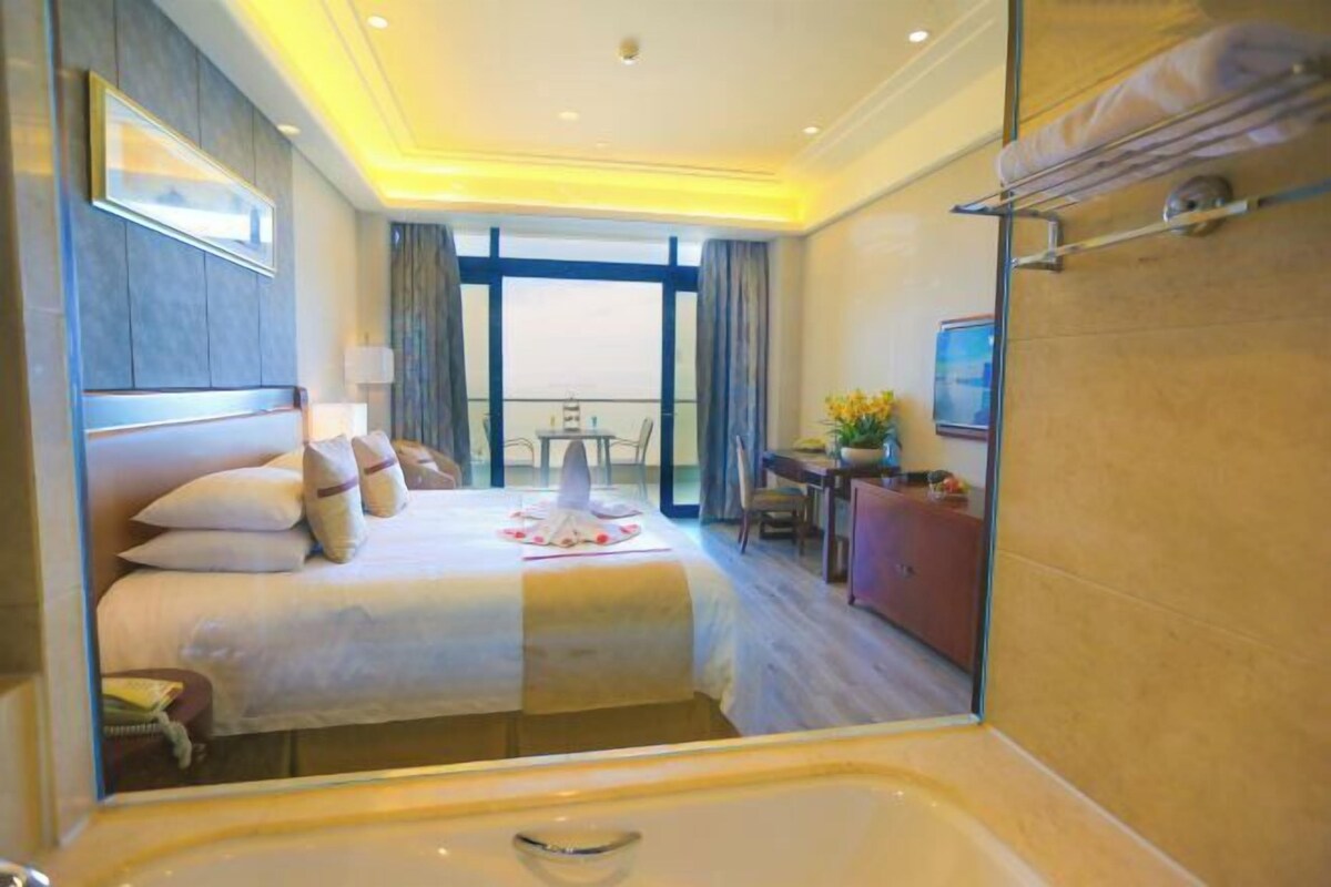 A hotel room with a large bed, a desk, a chair, a TV, and a balcony with a view.