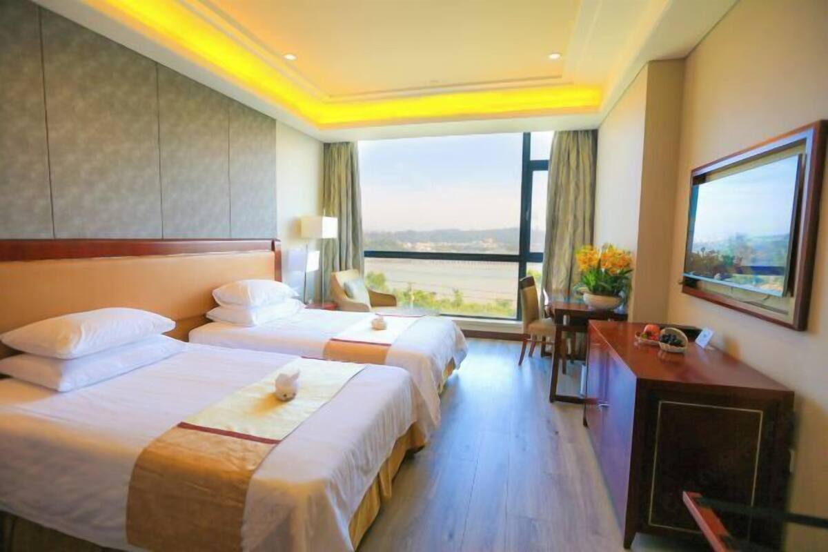 A hotel room with two beds, a desk, a chair, and a large window offering a view.