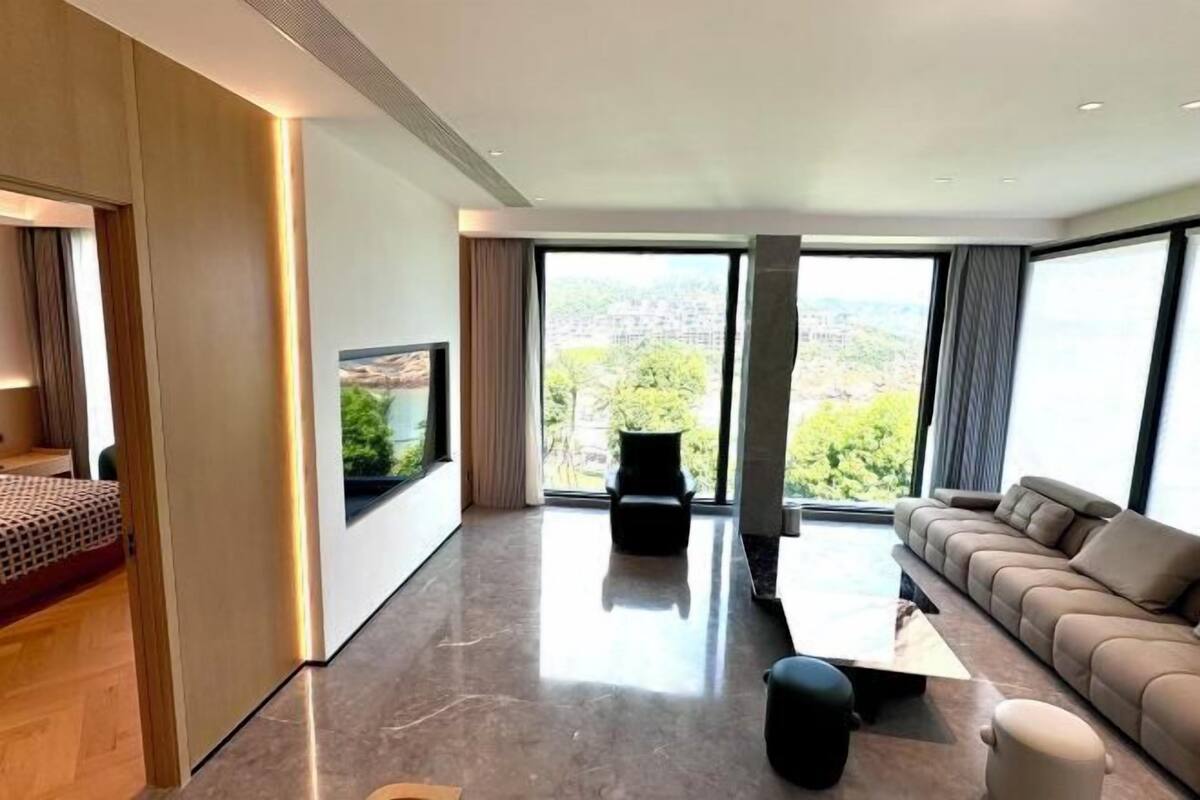A modern living room with a large window, a sofa, a coffee table, and a TV mounted on the wall.