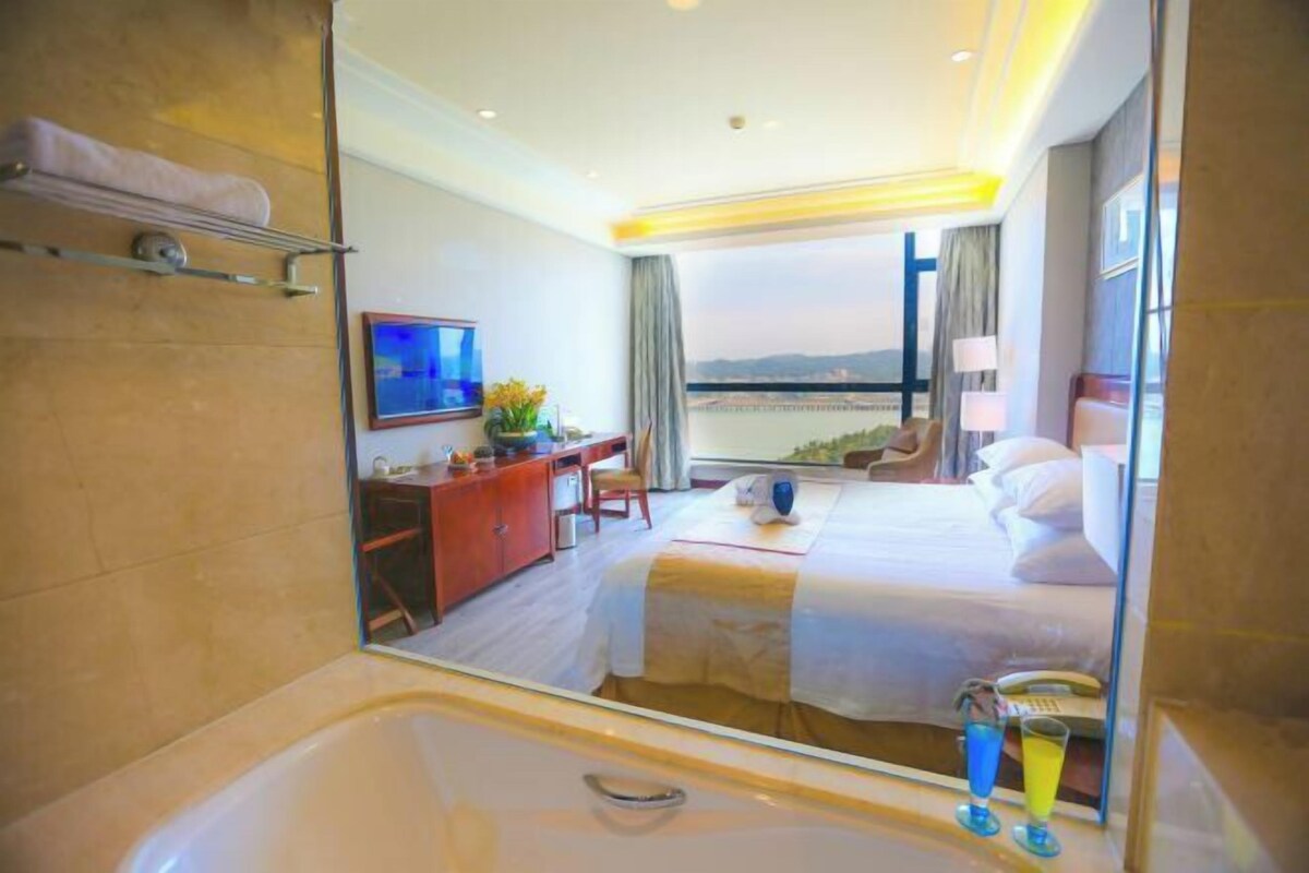 A hotel room with a large bed, a desk, a chair, a TV, a bathroom with a bathtub, and a view of the city.