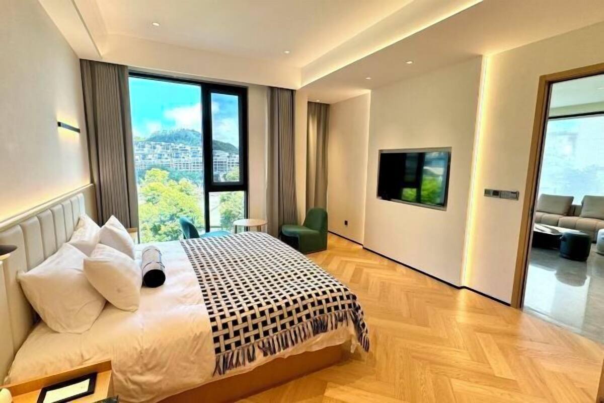 A modern hotel room with a large bed, a flat-screen TV, and a view of a cityscape through large windows.