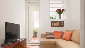 A living room with a TV on a wooden stand, a beige sofa with orange cushions, a checkered floor, a window with a cactus and flowers, and a door leading to a balcony with a potted plant. - 2 Classic Apartments by HostWise (Porto)
