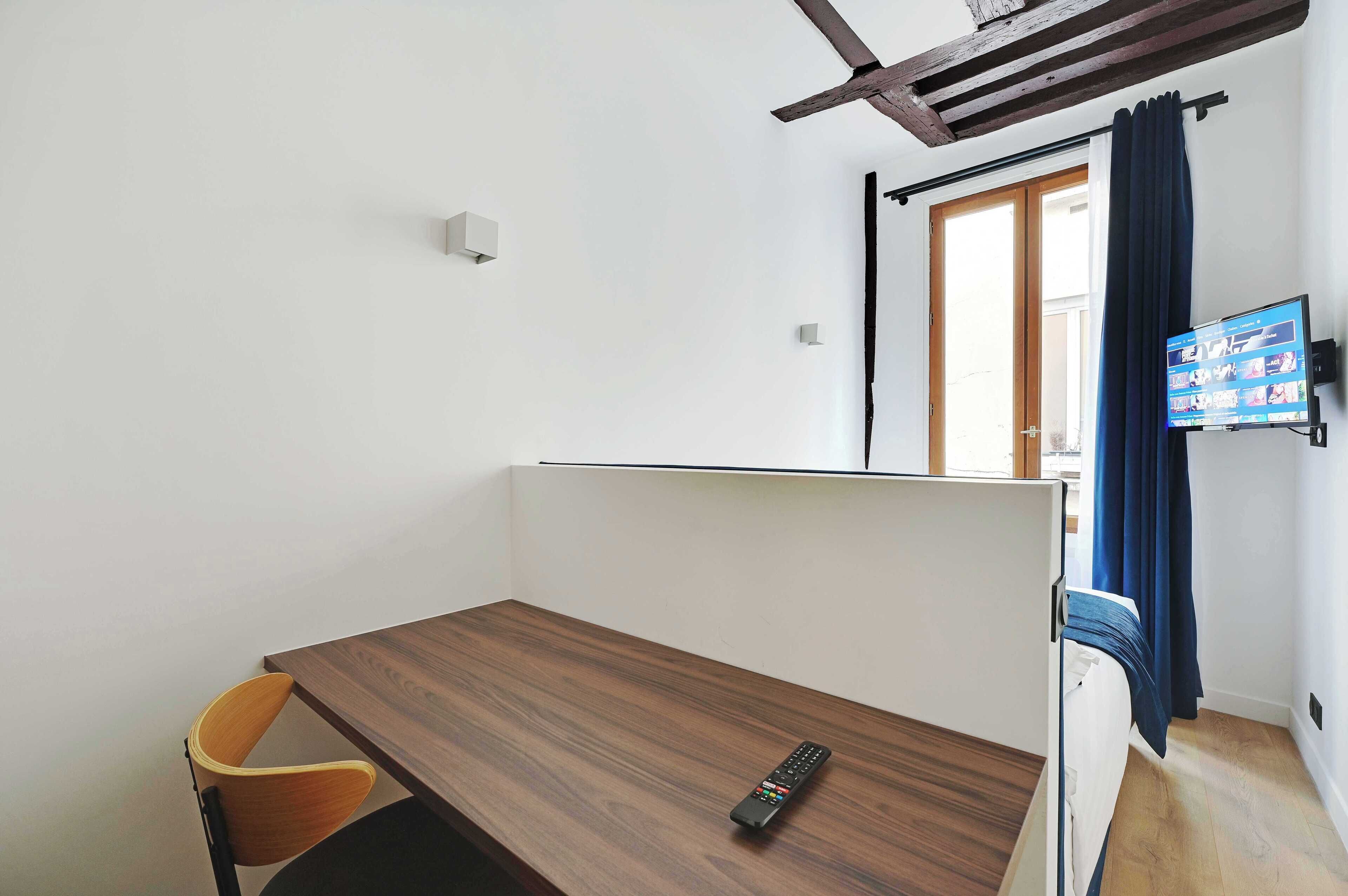 Une petite pièce moderne, avec un bureau en bois, une seule chaise, un téléviseur fixé au mur et une fenêtre avec des rideaux.