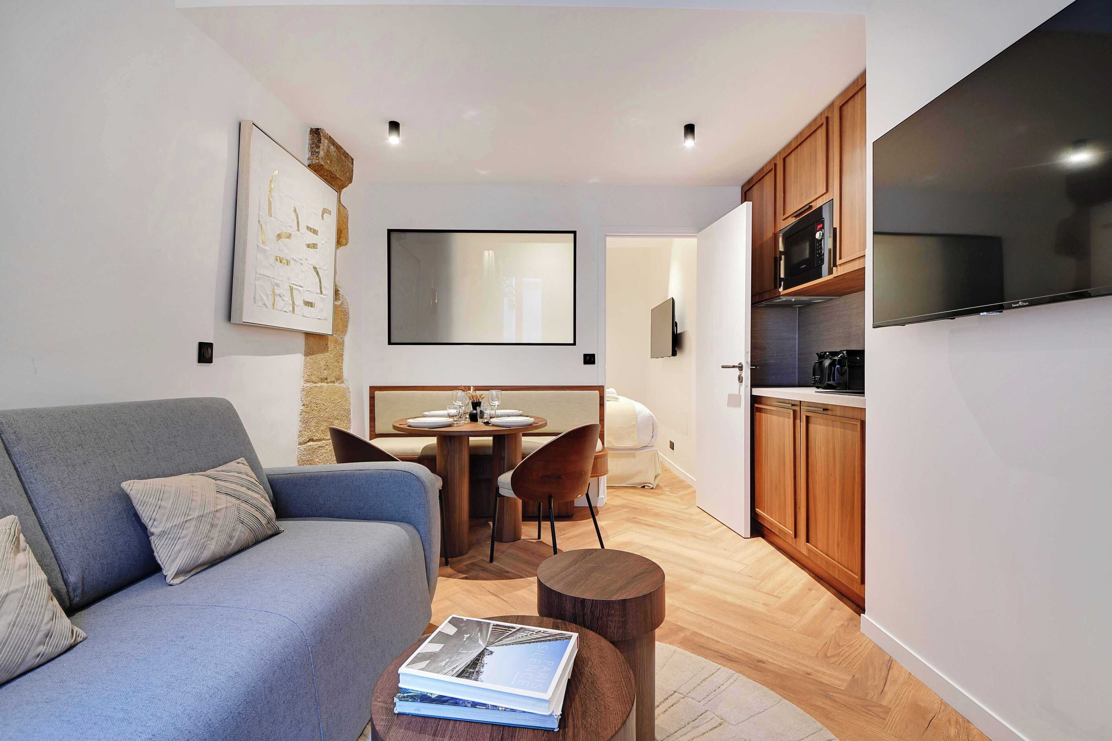 A modern living room with a sofa, a dining table, and a kitchen area with wooden cabinets and a flat-screen TV.