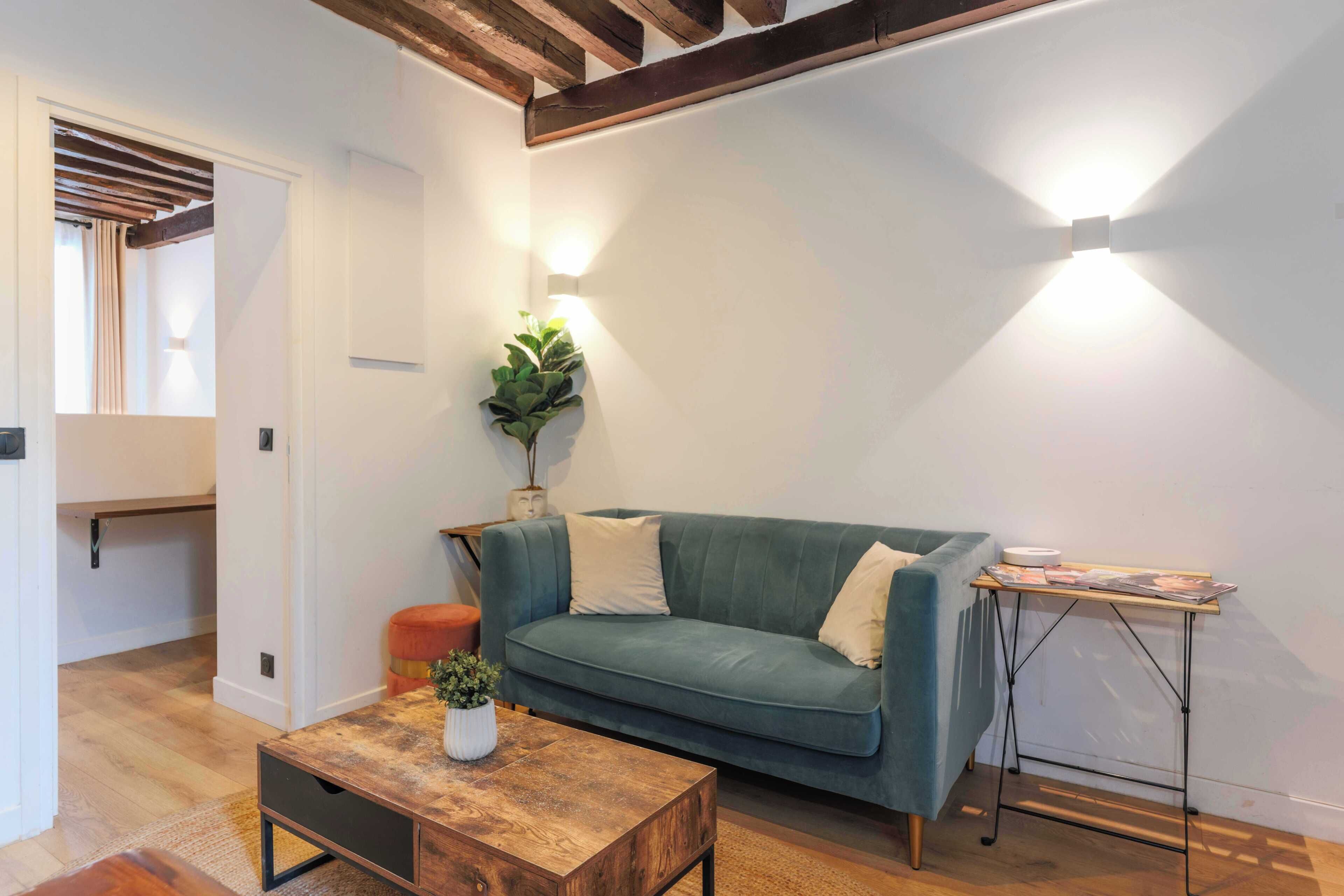 A living room with a teal sofa, a wooden coffee table, a small orange stool, a side table with a magazine, and a wall-mounted light fixture.