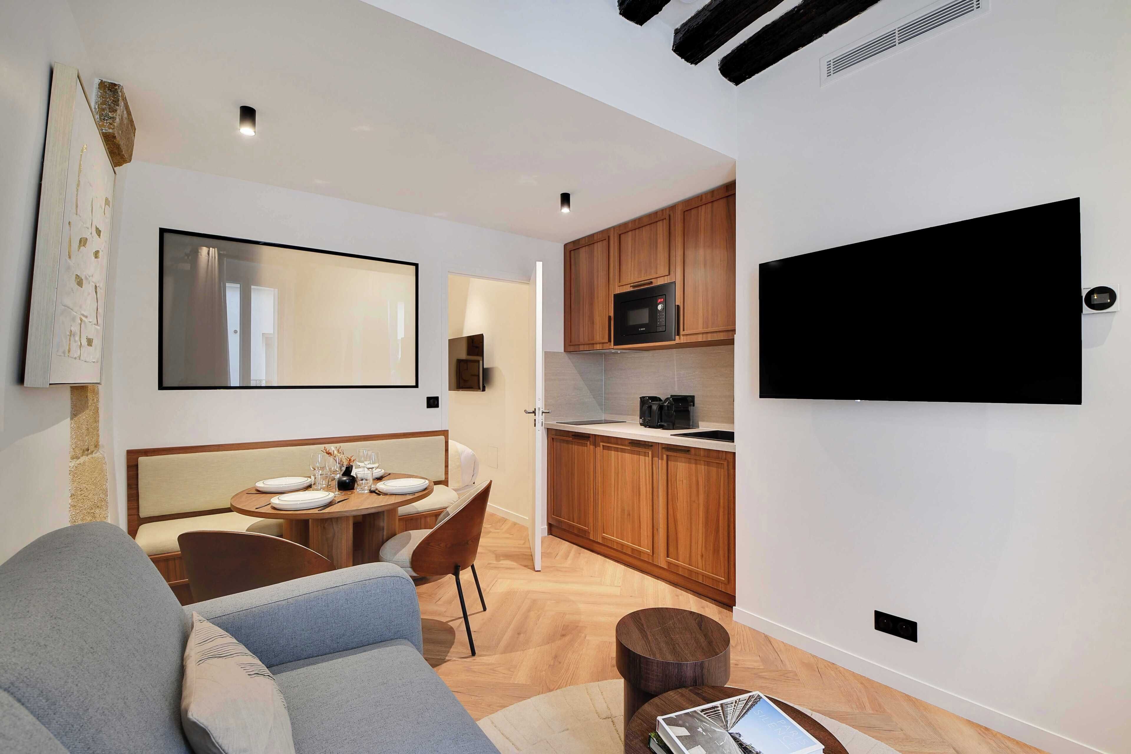 A modern living room with a sofa, a dining area with a table and chairs, a kitchen with wooden cabinets and a microwave, and a flat-screen TV mounted on the wall.