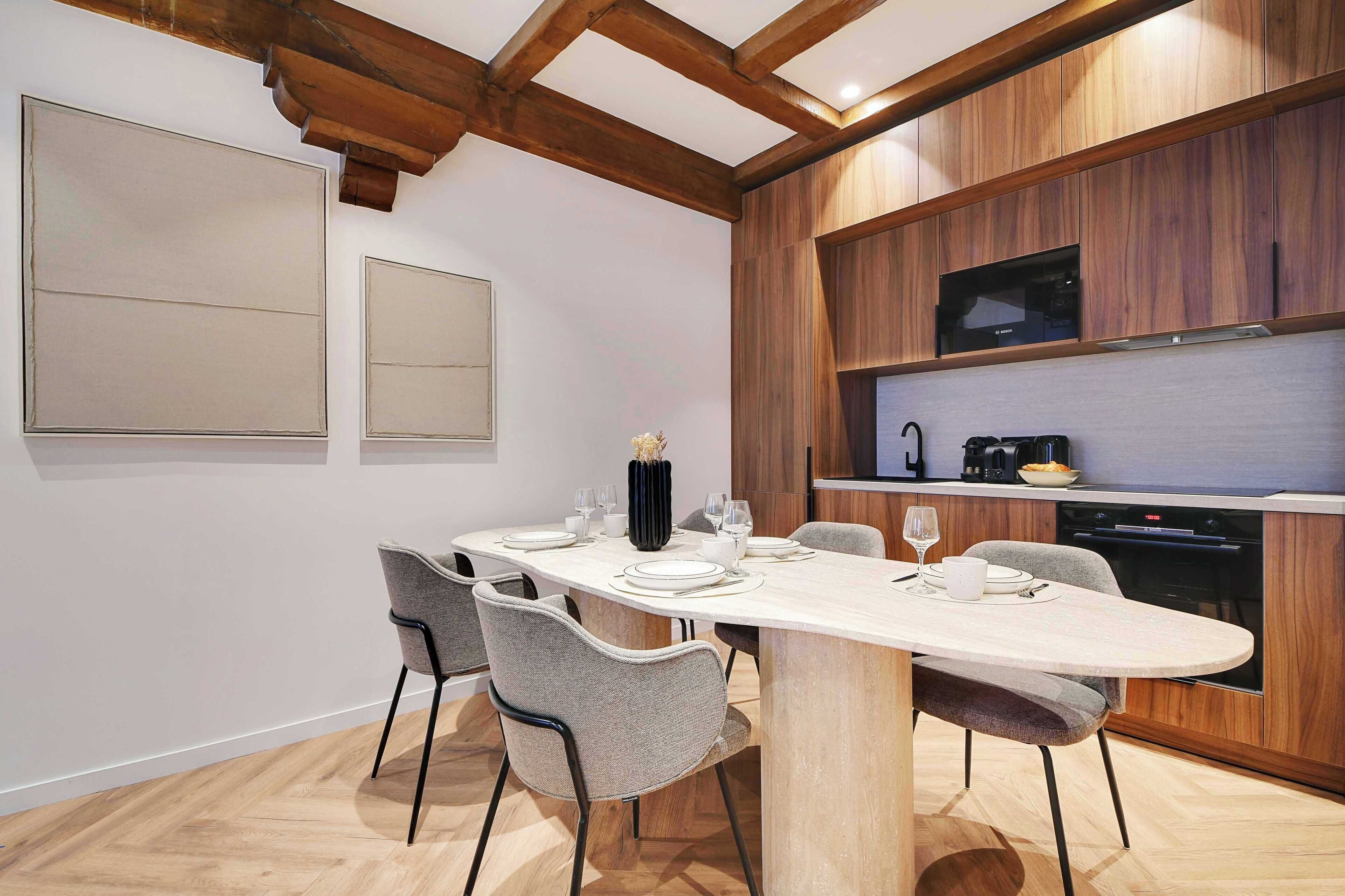 Een moderne eetruimte met een lange tafel voor vier personen, omringd door grijze stoelen. Een houten plafond met zichtbare balken en een ingebouwde magnetron boven het aanrecht is zichtbaar.