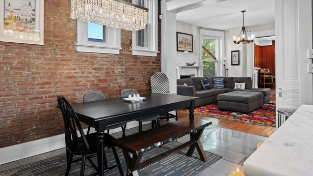 Ein modernes Wohnzimmer mit Backsteinwand, einer Essecke mit dunklem Holztisch und -stühlen sowie einem grauen Sofa.