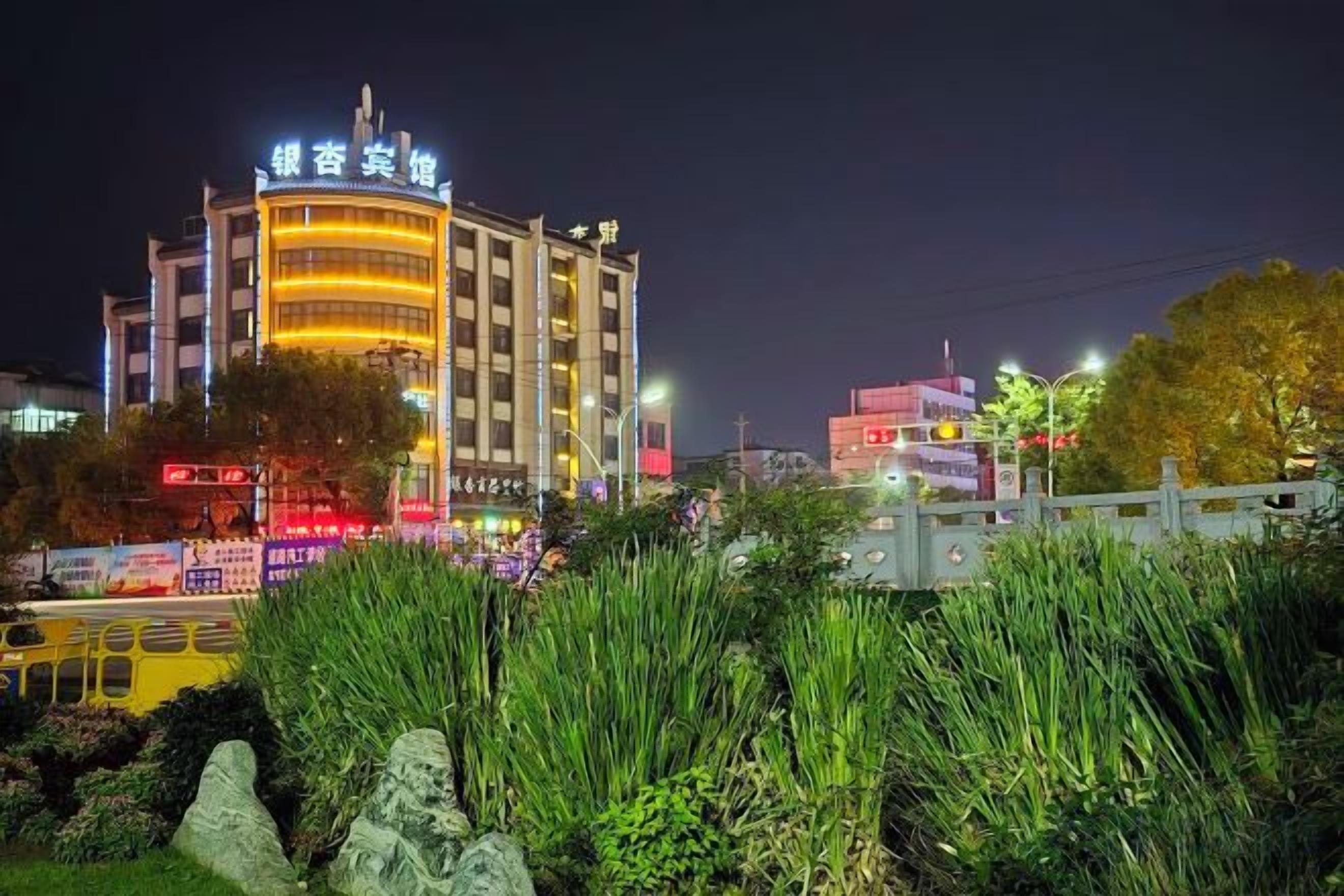 A multi-story building with illuminated signage, surrounded by greenery and a street with vehicles.